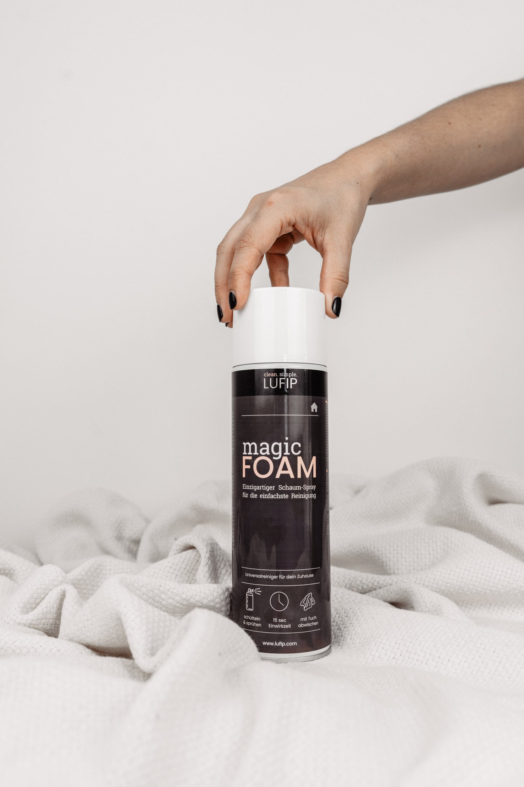 Magic Foam Schaumreiniger-Dose auf weißer Decke mit neutralem Hintergrund. Hand greift von oben rechts nach der Dose.