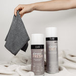 Auf einer weißen Decke vor einem neutralen Hintergrund liegt ein Set von Reinigungsprodukten. Von rechts greift eine Hand ins Bild, die ein gefaltetes Magic Towel hält. Das Set bietet vielseitige Reinigungslösungen für Schuhe und andere Oberflächen, mit dem Magic Towel als Teil dieses umfassenden Reinigungsangebots.
