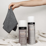 Auf einer weißen Decke vor einem neutralen Hintergrund liegt ein Set von Reinigungsprodukten. Von rechts greift eine Hand ins Bild, die ein gefaltetes Magic Towel hält. Das Set bietet vielseitige Reinigungslösungen für Schuhe und andere Oberflächen, mit dem Magic Towel als Teil dieses umfassenden Reinigungsangebots.