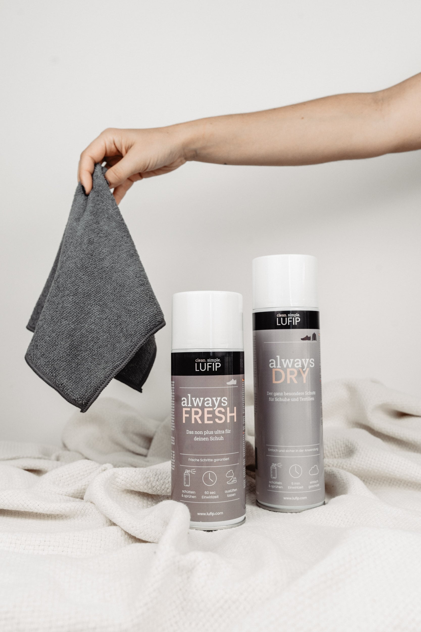 Auf einer weißen Decke vor einem neutralen Hintergrund liegt ein Set von Reinigungsprodukten. Von rechts greift eine Hand ins Bild, die ein gefaltetes Magic Towel hält. Das Set bietet vielseitige Reinigungslösungen für Schuhe und andere Oberflächen, mit dem Magic Towel als Teil dieses umfassenden Reinigungsangebots.