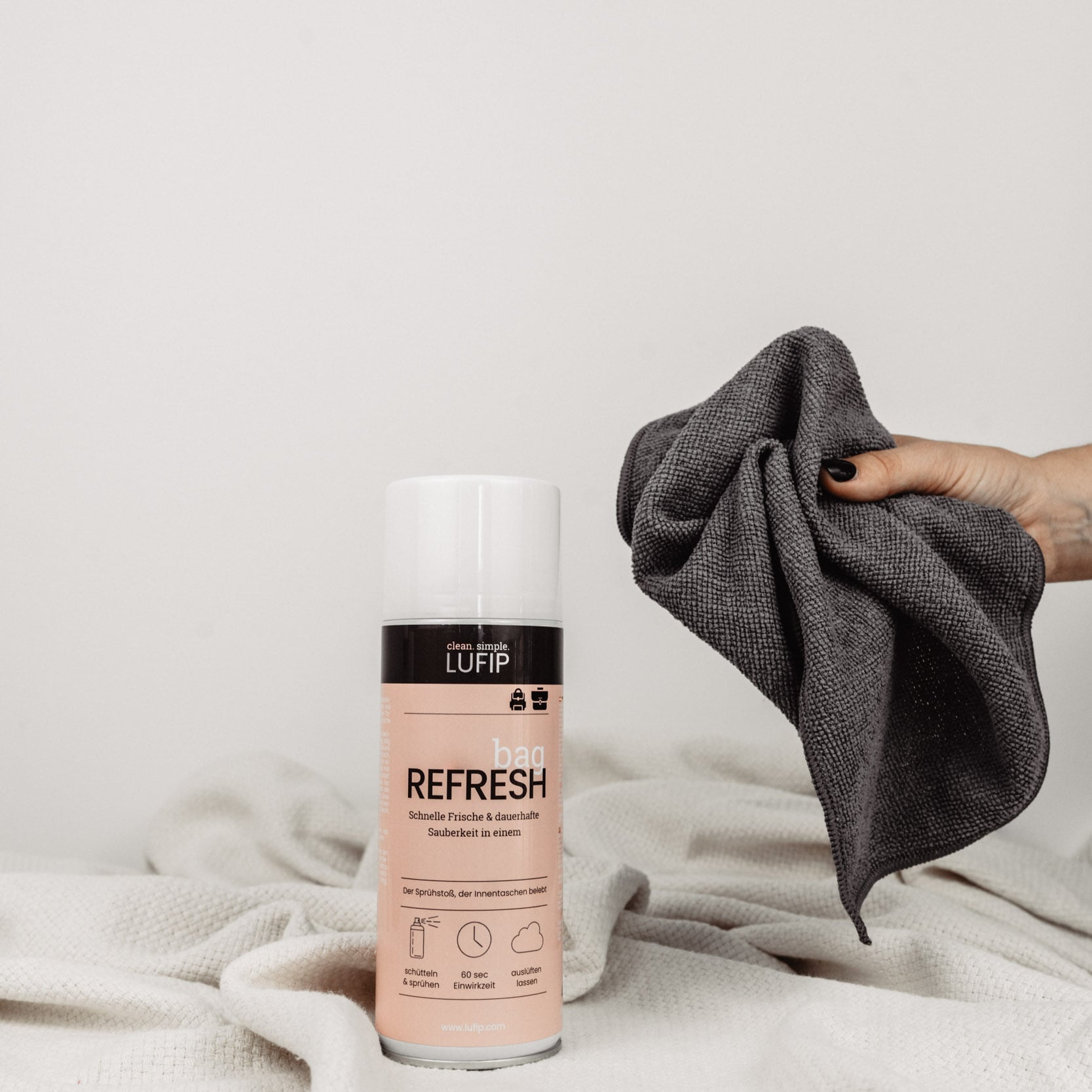 Bag Refresh auf weißer Decke vor neutralem Hintergrund. Hand greift von rechts ins Bild mit Micro Towel.