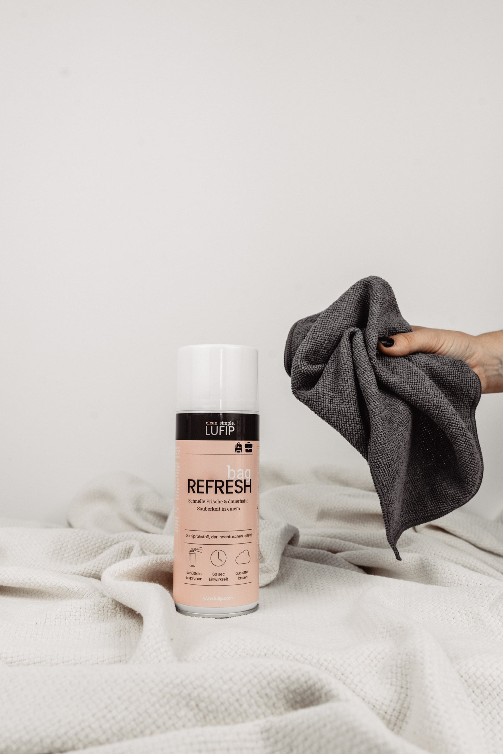 Bag Refresh auf weißer Decke vor neutralem Hintergrund. Hand greift von rechts ins Bild mit Micro Towel.