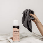 Bag Refresh auf weißer Decke vor neutralem Hintergrund. Eine Hand greift von rechts ins Bild mit dem Micro Towel.