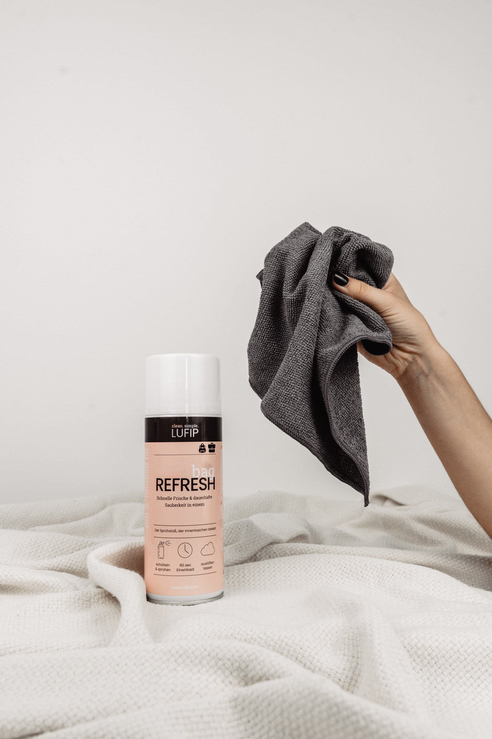 Bag Refresh auf weißer Decke vor neutralem Hintergrund. Eine Hand greift von rechts ins Bild mit dem Micro Towel.